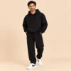 Man wearing the Sportlux Plus black 350 GSM fleece hoodie co-ord set, relaxed hoodie and straight leg trousers with tonal embroidered S monogram, full length view