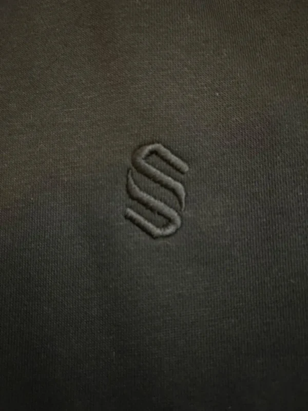 Close-up of the tonal black embroidered S monogram on the Sportlux Plus black fleece hoodie