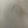 Close-up of the tonal cream embroidered S monogram on the Sportlux Plus cream fleece hoodie